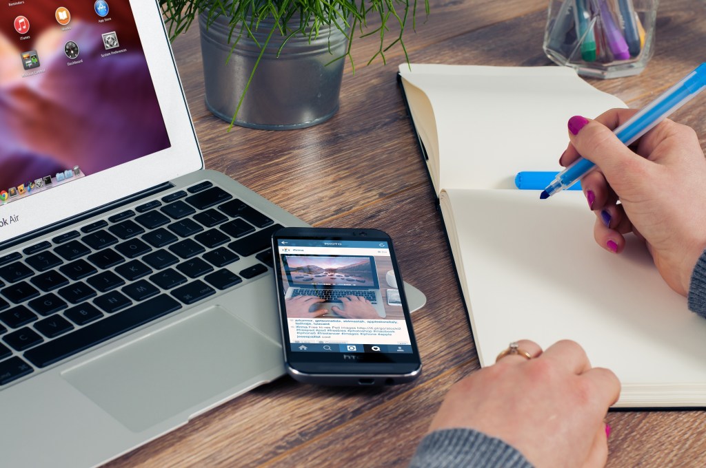 picture of a laptop, phone, and journal. A hand is holding a pen ready to write.